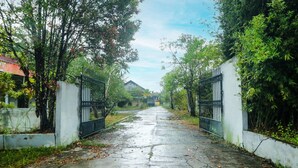A concrete pathway flanked by metal gates and lush greenery, leading to a building with a traditional roof. - Hotel O Nyi Rindang Near Puskesmas Jekan Raya (Palangkaraya)