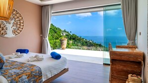 Un dormitorio con cama, toallas, una mesita de noche de madera y vistas al océano y la vegetación.