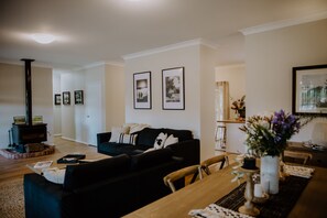 Een woonkamer met een zwarte bank, een houten eettafel en een open haard. Er hangen ingelijste foto's aan de muur en staat een vaas met bloemen op de tafel.