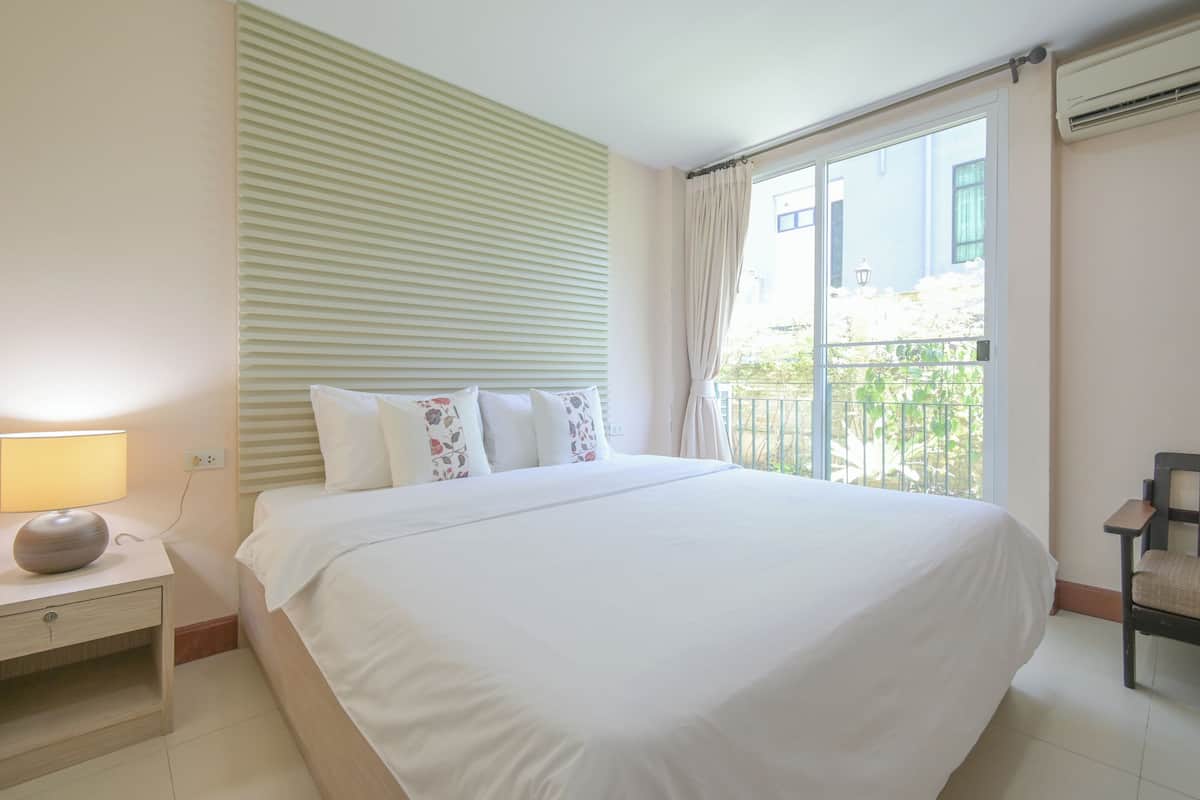 A hotel room with a large bed, a bedside table with a lamp, a chair, and a window with a view of greenery.