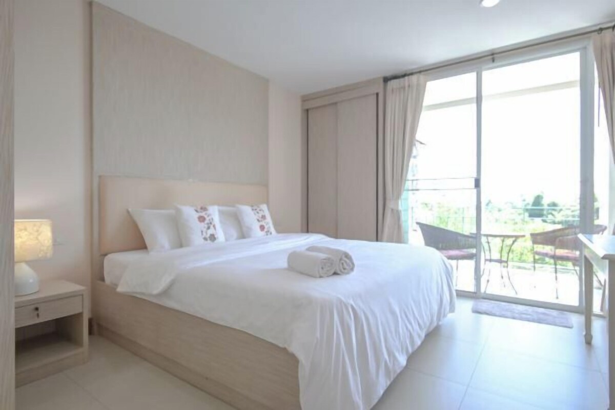 A hotel room with a large bed, a nightstand, a window with curtains, and a view of the outdoors.