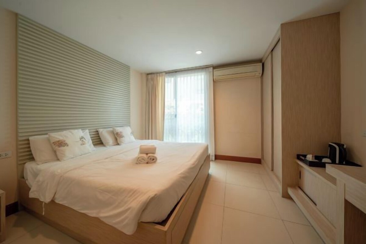 A hotel room with a large bed, a wooden wardrobe, a small bedside table, and a window with curtains.