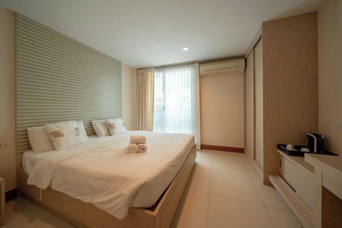 A hotel room with a large bed, a wooden wardrobe, a small bedside table, and a window with curtains.