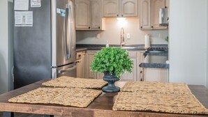 A kitchen with wooden cabinets, a stainless steel refrigerator, a potted plant, and woven placemats on the table. - Charming KY Lake Condo Short Walk to Lake (Gilbertsville)