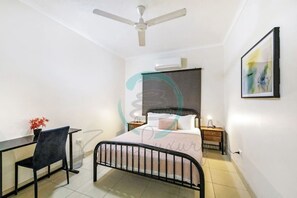 Une chambre à coucher comprenant un lit, un bureau, une chaise, un ventilateur de plafond et un climatiseur fixé au mur.
