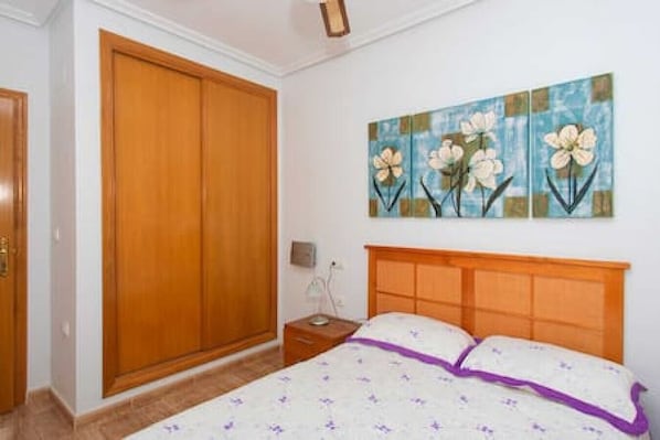 A bedroom with a wooden wardrobe, a bed with a purple accent, and a floral painting on the wall. - CH Isamar Park Doña Pepa (Ciudad Quesada) (Ciudad Quesada)