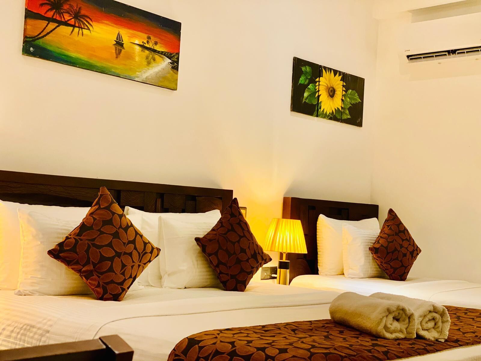 A hotel room with a bed, pillows, a lamp, and two paintings on the wall.