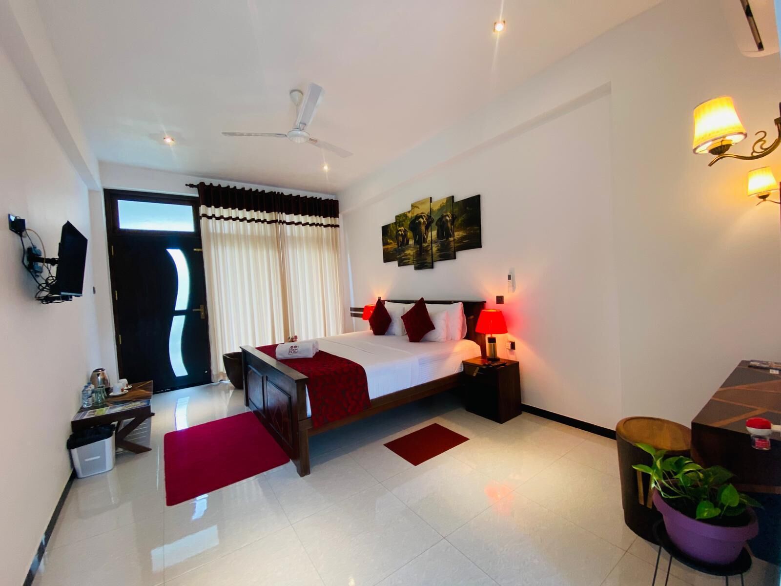 A hotel room with a bed, bedside tables, a desk, a chair, a TV, a ceiling fan, a window with curtains, and a wall-mounted artwork.
