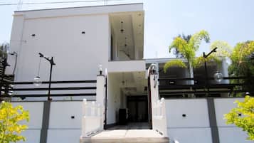 A modern building with a white facade, a central entrance with a glass door, and a black railing.