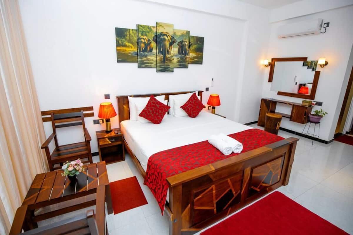 Ein Hotelzimmer mit einem hölzernen Bett, rot-weiß gestreifter Bettwäsche, einem Schreibtisch mit Stuhl, einem kleinen Tisch mit Blumenvase und einem an der Wand befestigten Spiegel.