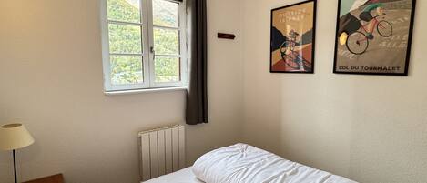 Un dormitorio con cama, ventana con cortinas, radiador, lámpara y dos cuadros en la pared.