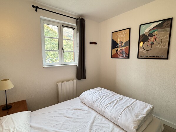 A bedroom with a bed, a window with curtains, a radiator, a lamp, and two framed pictures on the wall. - Duplex + terrasse panoramique.Parking.Piscine été (Cauterets)