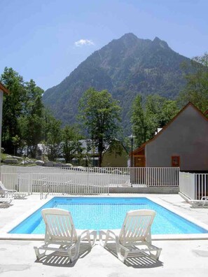 A swimming pool with two lounge chairs in the foreground, surrounded by a white fence. In the background, there is a mountain and a building with a red roof.