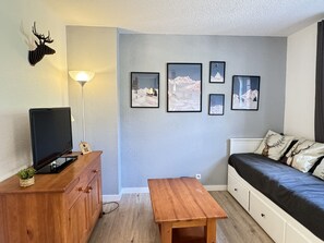 Una habitación pequeña y acogedora con una cama, un televisor sobre un mueble de madera, una mesita y un panel decorado con marcos.