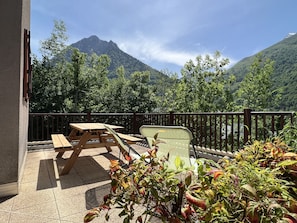 A balcony with a wooden table and chairs, surrounded by plants, overlooking a mountainous landscape.