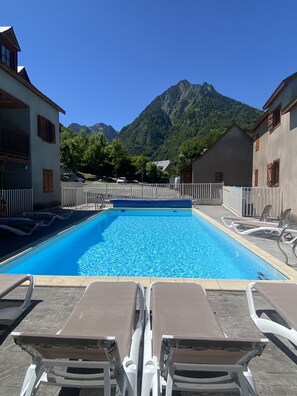 Piscina com espreguiçadeiras, rodeada por edifícios e com vista para uma montanha.