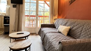 A cozy living room with a sofa, a small round table, a TV mounted on the wall, and a balcony with a view of the outdoors. - T3 rénové avec balcon, vue montagne et Parking. (Cauterets)