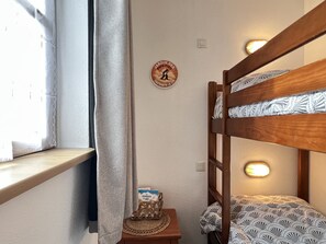 A bunk bed with a patterned bedspread, a small table with a decorative item, a wall clock, and a window with a curtain. - T3 rénové avec balcon, vue montagne et Parking. (Cauterets)