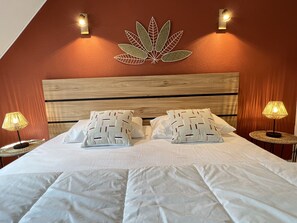 Cama bem arrumada com lençóis brancos e almofadas decorativas, cabeceira de madeira e duas luminárias fixadas na parede.
