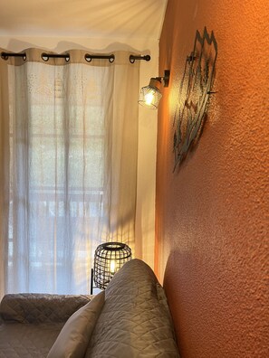 A room with a bed, a wall-mounted light fixture, and a window with sheer curtains.