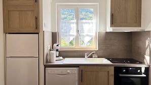 A compact kitchen with a dishwasher, stove, and a window with a view of greenery. - T3 rénové avec balcon, vue montagne et Parking. (Cauterets)