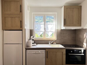 A compact kitchen with a dishwasher, stove, and a window with a view of greenery. - T3 rénové avec balcon, vue montagne et Parking. (Cauterets)