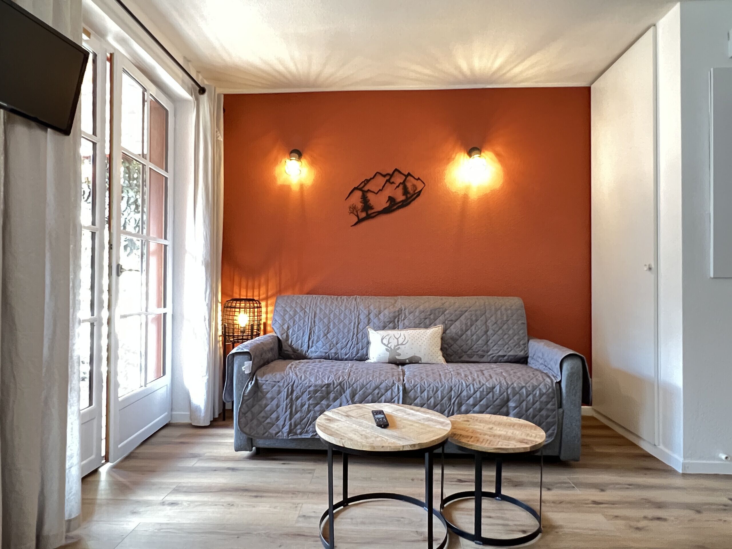 Une pièce avec un canapé gris, deux lampes fixées au mur, un élément décoratif mural et une petite table ronde sur laquelle se trouve une télécommande.