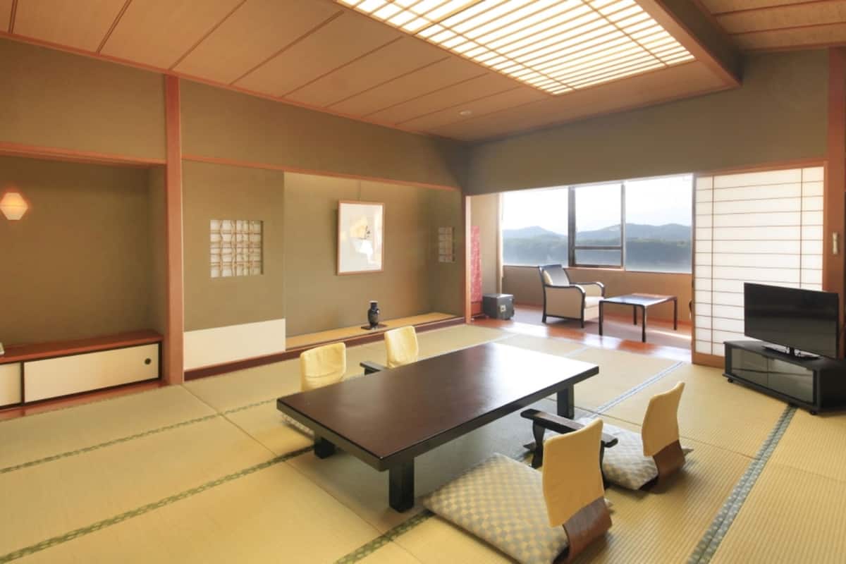 A traditional Japanese-style room with tatami flooring, a low table, chairs, a television, and a view of mountains through large windows.