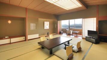 A traditional Japanese-style room with tatami flooring, a low table, chairs, a television, and a view of mountains through large windows.