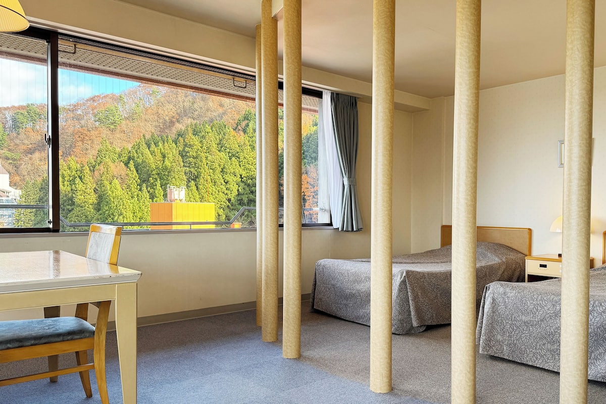 A hotel room with two beds, a desk, and a window with a view of trees and buildings.