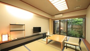 Ein traditioneller japanischer Raum mit Tatami-Boden, einem niedrigen Tisch, einem Fernseher und einer Schiebeglastür zu einem Garten.