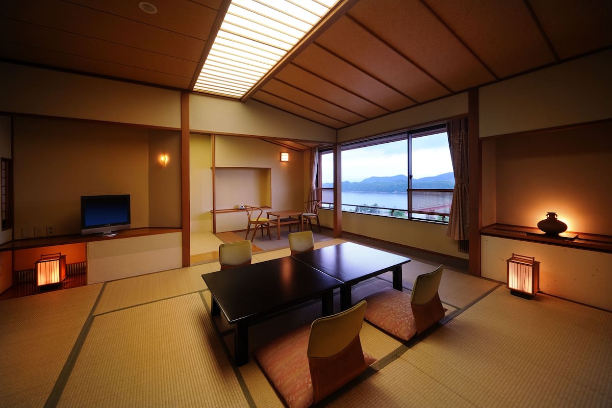 A traditional Japanese room with tatami flooring, a low table, and chairs. A television is on a low stand, and there is a large window with a view of mountains.