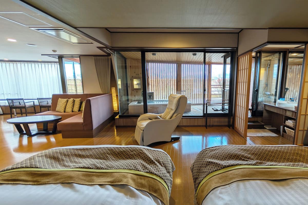 A hotel room with a sofa, armchair, two beds, a desk, and a sliding glass door to a balcony.