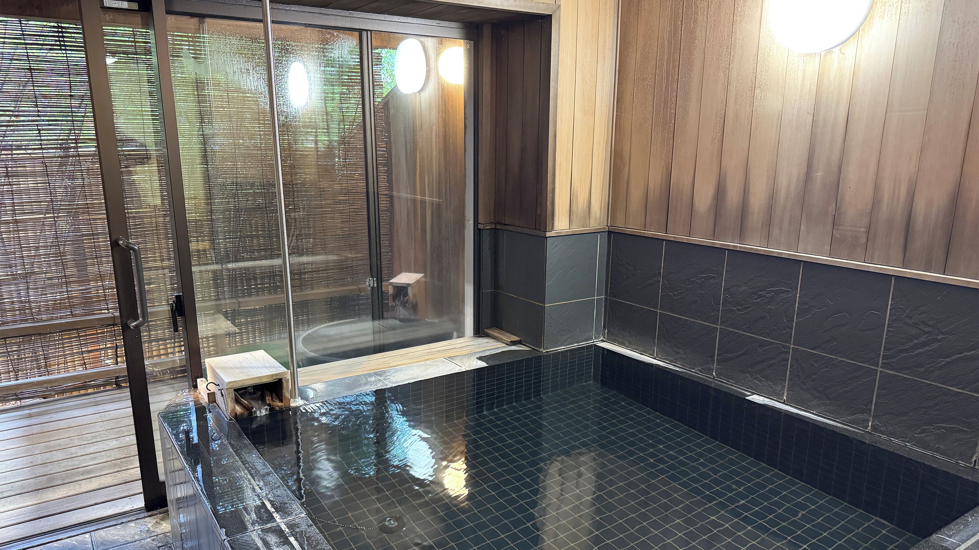 Un baño moderno con cabina de ducha de vidrio, paredes de azulejos oscuros y una terraza de madera en el exterior.