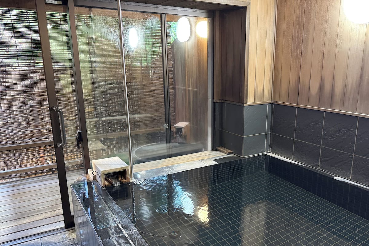 A modern bathroom with a glass shower enclosure, dark tiled walls, and a wooden deck outside.