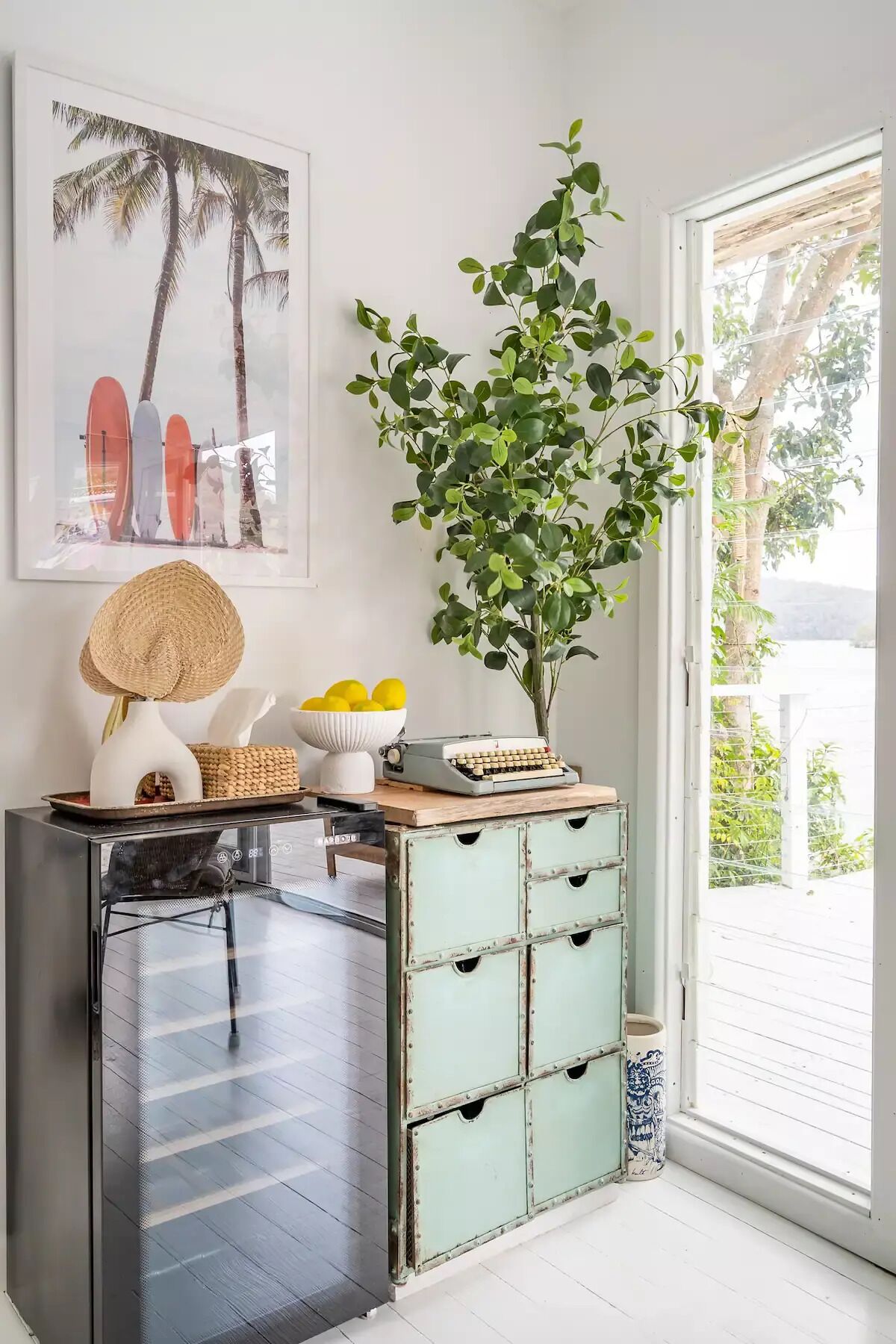 A room with a glass door leading to a balcony, a vintage typewriter, a woven basket, a bowl of lemons, and a framed picture of surfboards.