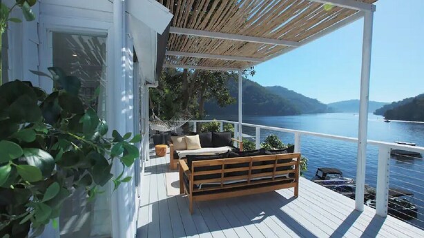 Una terraza de madera con un banco y cojines, con vistas a un lago tranquilo con barcos y colinas al fondo.