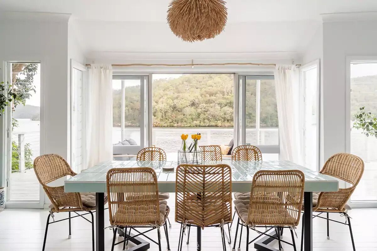 A dining area with a glass table surrounded by wicker chairs, large windows with a view of trees, and a hanging light fixture.