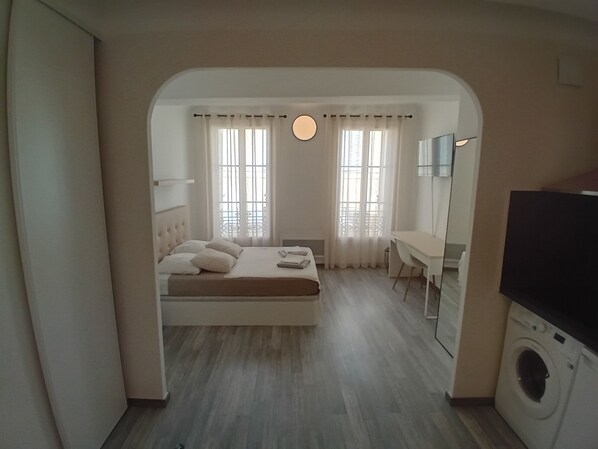 A bedroom with a bed, a desk, a washing machine, and a large window with curtains.