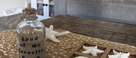 Lasinen pullo, jonka etiketissä lukee ”Lempirantani hiekka”, on asetettu punotulle alustalle. Sen ympärillä on puutarjolla meritähtikuviolla koristeltuja keksejä.