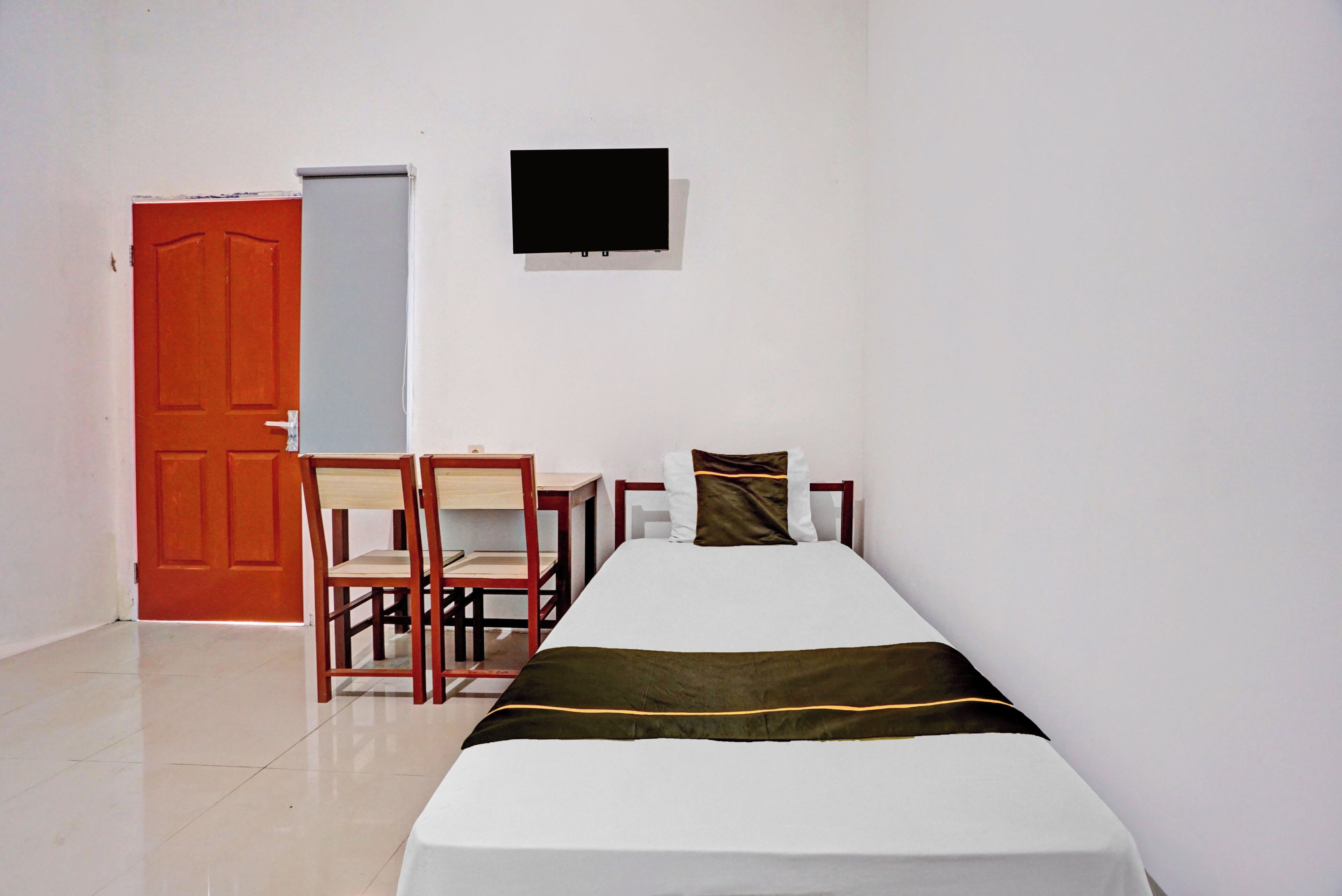 A single bed with a white and brown bedspread, a desk with a chair, a television mounted on the wall, and an orange door.