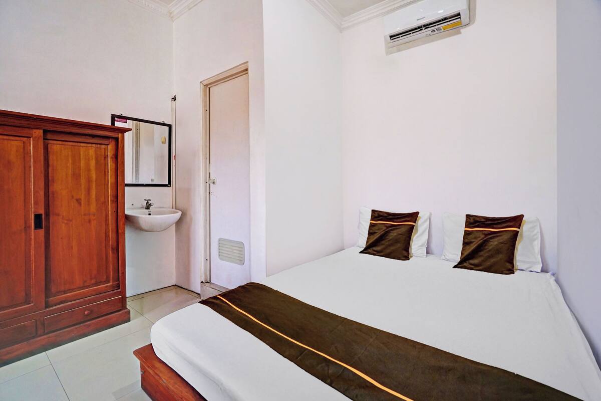 Habitación de hotel con armario de madera, lavamanos blanco, cama con ropa de cama blanca y marrón, y equipo de aire acondicionado.
