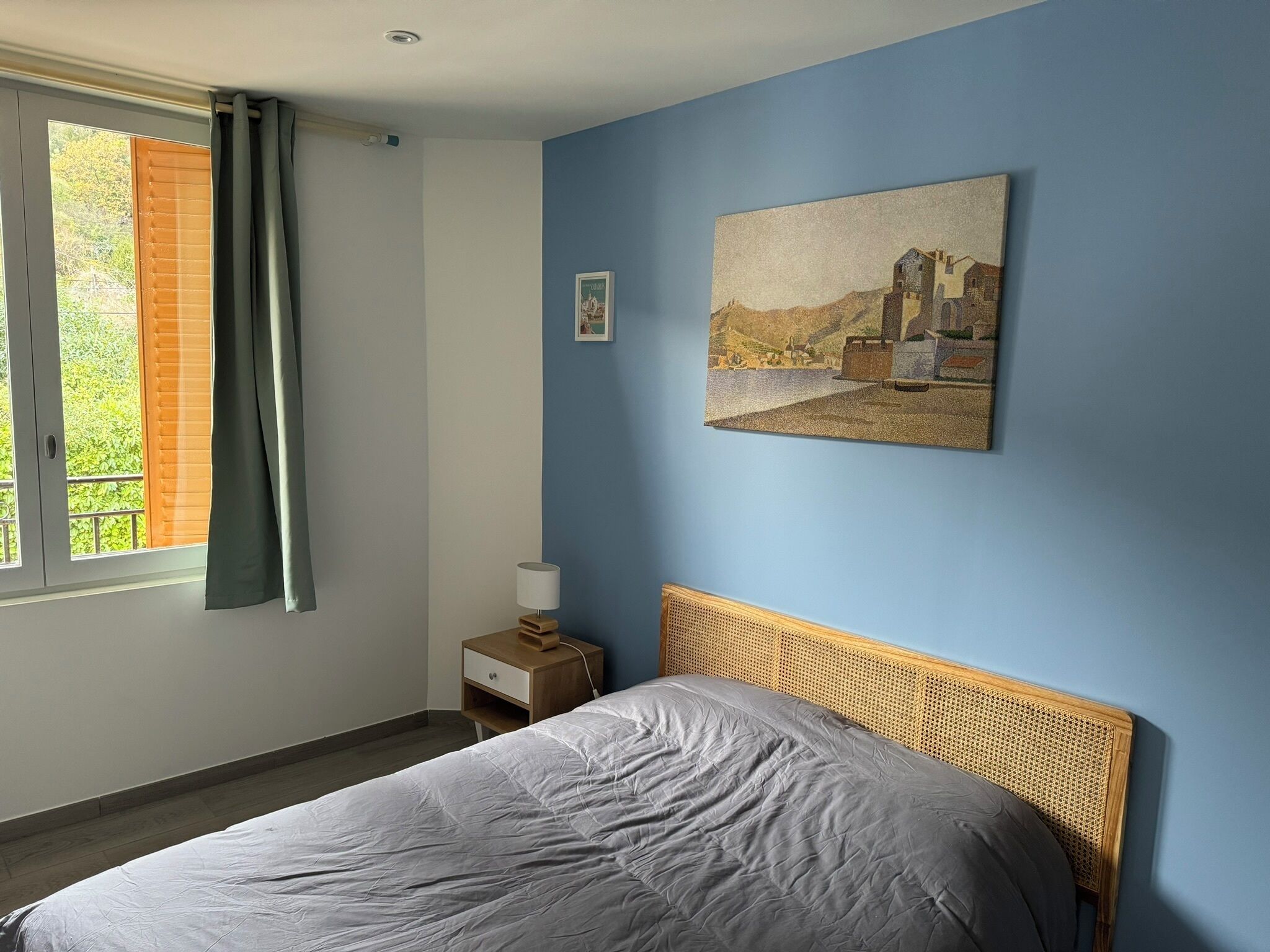 A bedroom with a bed, a nightstand, a lamp, a painting, and a window with curtains.