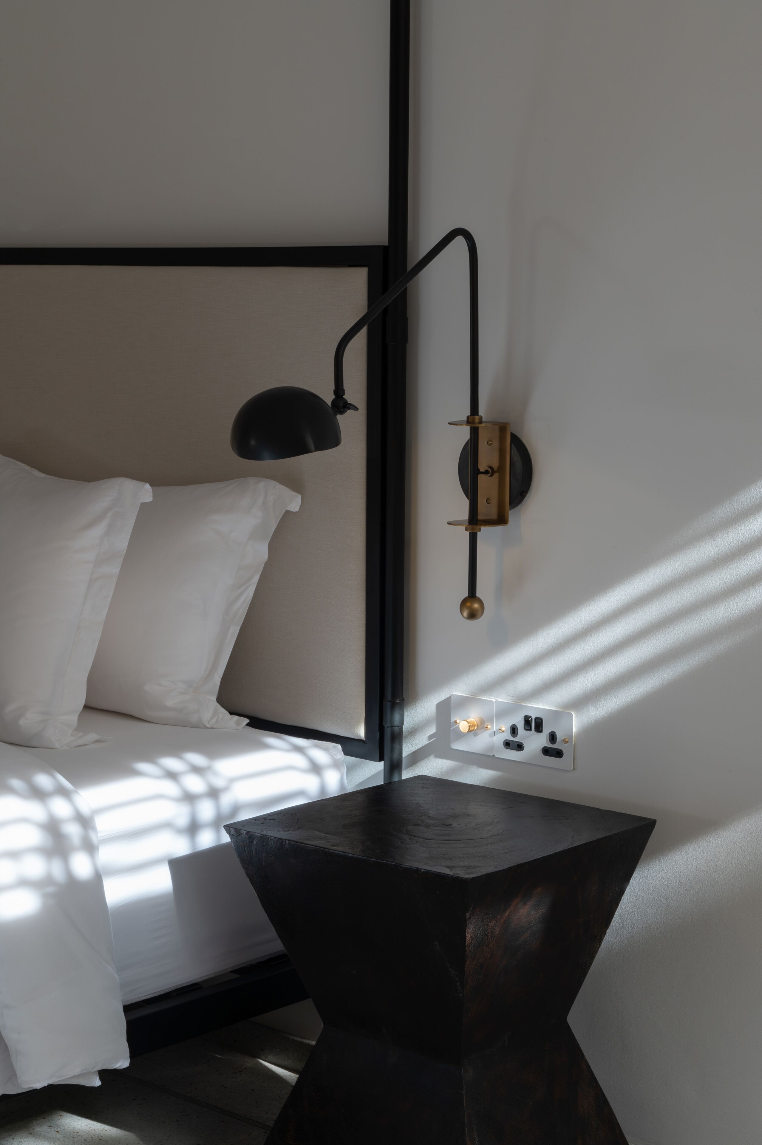 Una cama con sábanas blancas, una mesita de noche negra y una lámpara de pared.