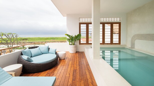 Ein moderner Poolbereich im Freien mit einer Holzterrasse, einer runden Sitzecke mit Kissen und Blick aufs Meer.