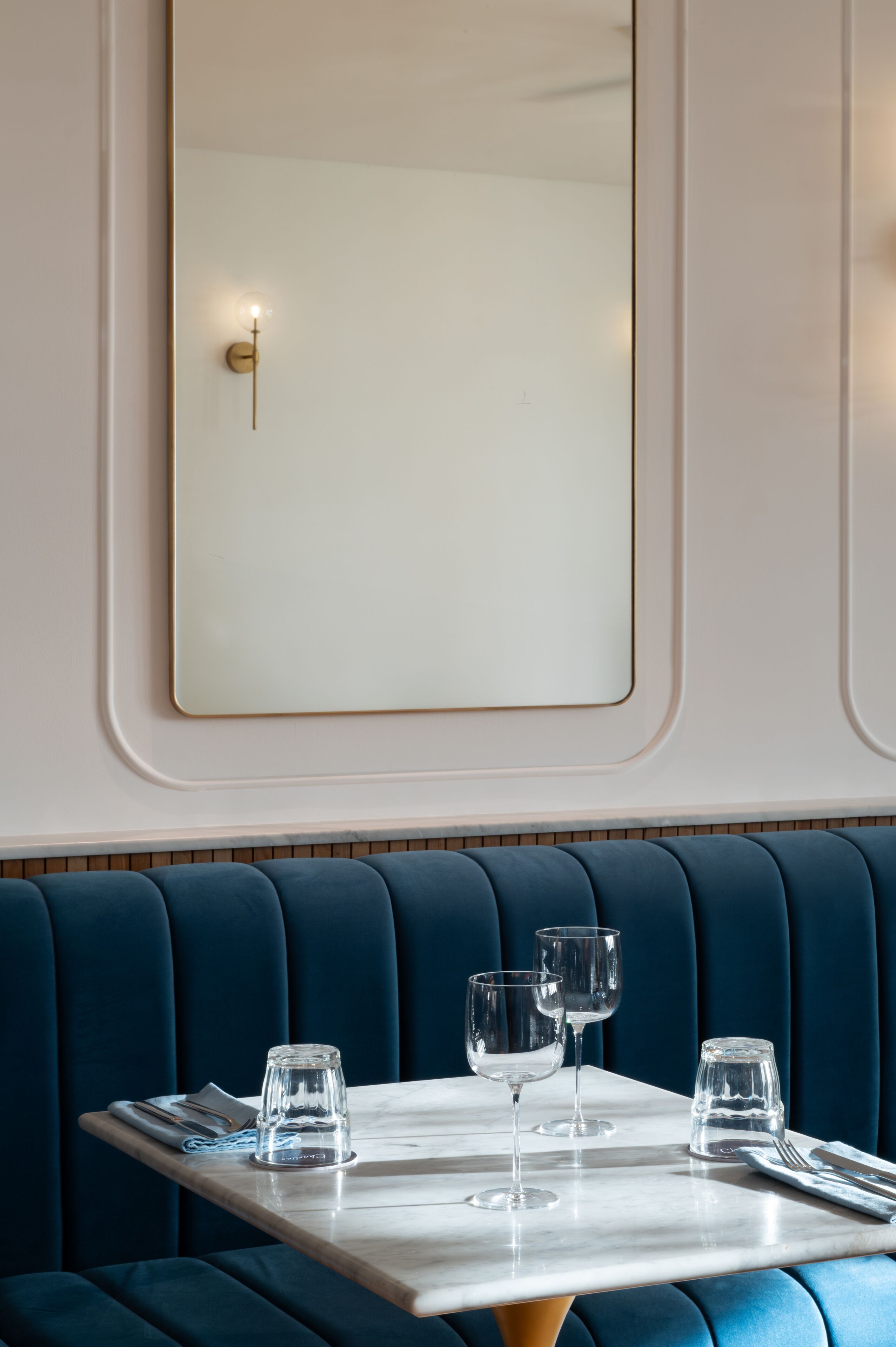 Un comedor con una mesa dispuesta para dos personas, un asiento tapizado azul y un espejo montado en la pared.