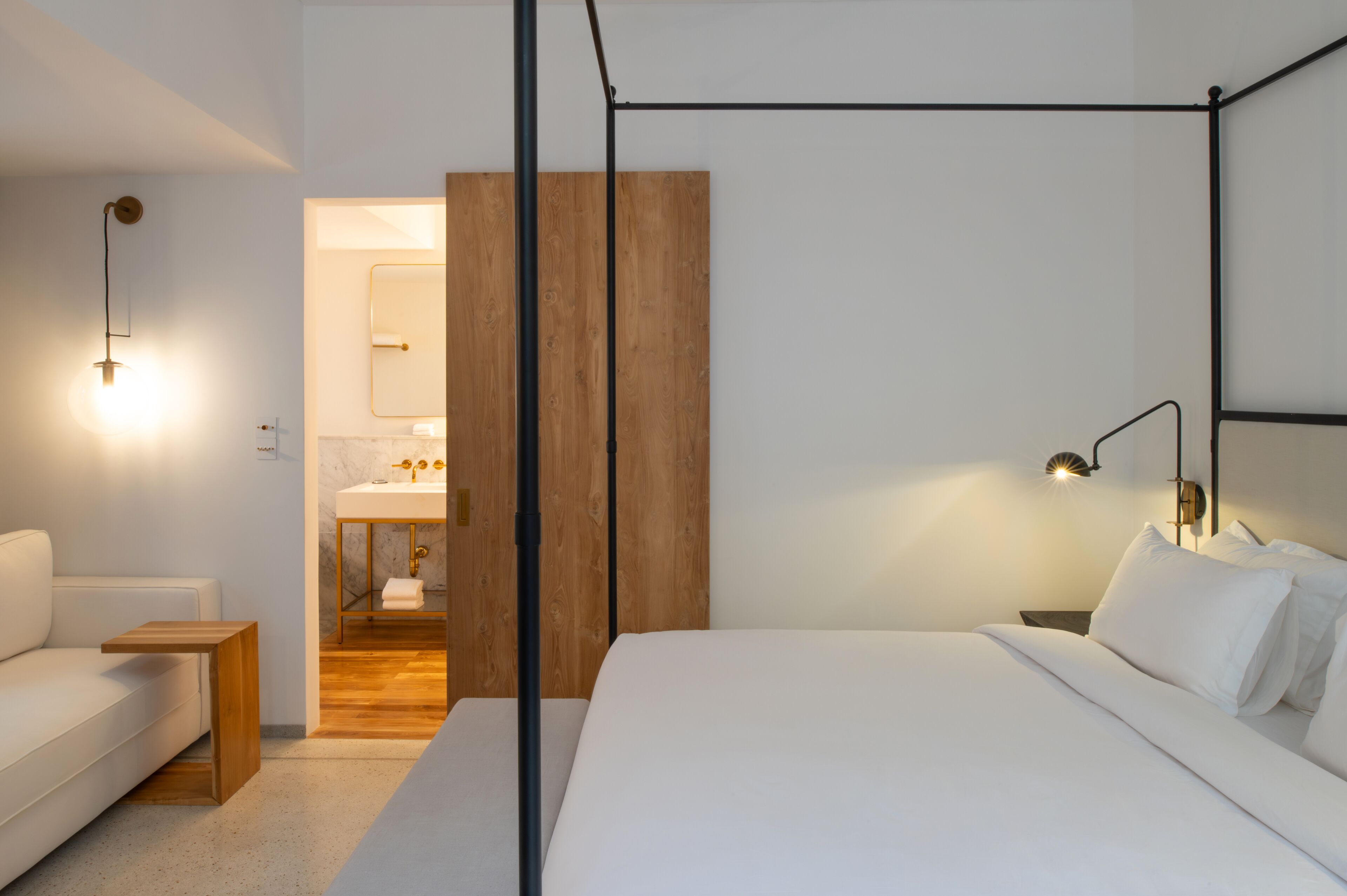 A modern bedroom with a four-poster bed, a wooden bedside table, a white sofa, and a bathroom with a wooden vanity and a mirror.