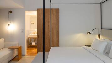 A modern bedroom with a four-poster bed, a wooden bedside table, a white sofa, and a bathroom with a wooden vanity and a mirror.