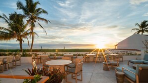 Eine Terrasse mit runden Tischen und Stühlen, Palmen und Blick auf den Sonnenuntergang über dem Meer.