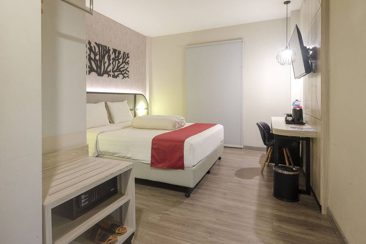 A hotel room with a bed, bedside table, desk, chair, and a trash bin.